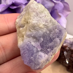 May include: A rough-cut, natural stone with a blend of lavender and cream hues. The stone's surface has a textured appearance, with areas of deep purple and lighter, almost white, sections. The stone is held in a hand, with a blurred purple flower in the background.