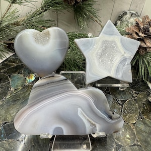 May include: Three agate slices in the shapes of a heart, star, and cloud. The agate is a light gray color with white and brown banding. The slices are displayed on clear acrylic stands.