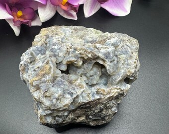 May include: A rough-hewn, natural stone specimen with a cavity, showcasing a mix of gray, blue, and beige hues. The stone's surface has a textured appearance. Purple orchid flowers are in the background.