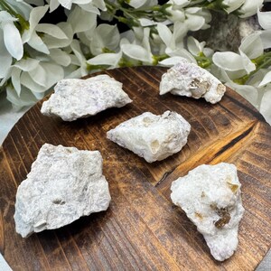 May include: Five raw, light-colored mineral specimens with subtle pink and brown hues are arranged on a round, dark wooden tray. White flowers are in the background. The minerals have a rough, textured appearance.