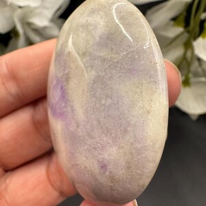 May include: A smooth, oval-shaped stone with a polished surface. The stone features a blend of colors, including lavender, gray, and cream. The stone is held in a hand, with white flowers in the background.