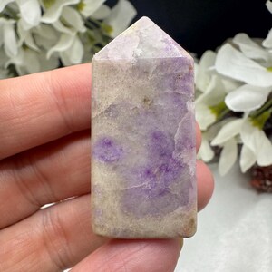 May include: A rectangular crystal tower with a pointed top. The stone features a blend of purple, lavender, and beige hues. The crystal is held in a hand, with white flowers in the background.