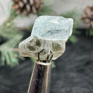 May include: A light blue aquamarine crystal with a clear, glassy texture. The crystal is mounted on a gray, textured base.