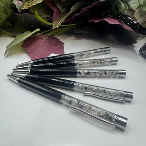 May include: A collection of black and silver pens with clear barrels filled with small, clear stones. The pens are arranged on a white surface, with green and purple foliage in the background. The pens have silver clips and tips.