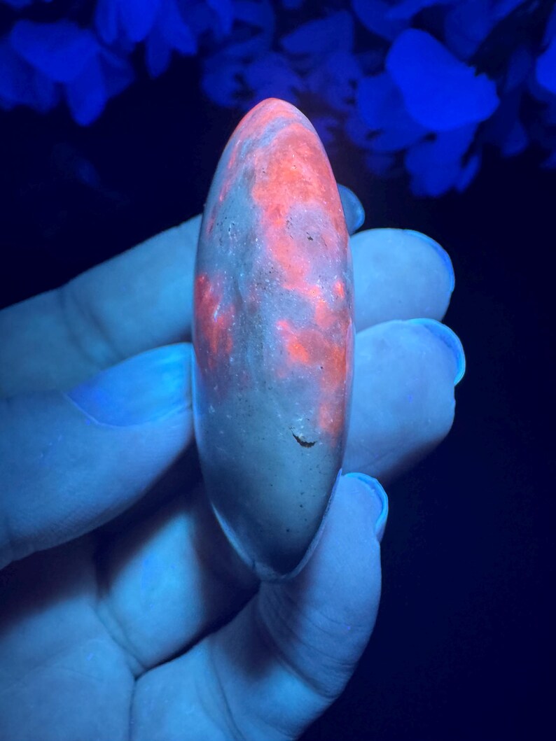 May include: A glowing, elongated stone with a speckled, reddish-orange pattern. The stone is held against a dark background, illuminated by a blue light. The stone's surface appears smooth with a slight curve.