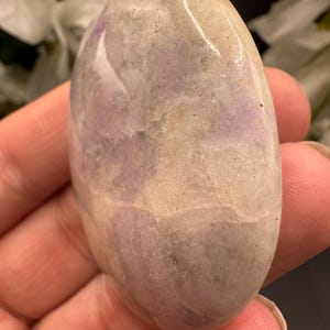 May include: Oval-shaped polished stone with a smooth, glossy surface. The stone features a blend of colors, including lavender, cream, and gray, with subtle variations in tone. The stone is held in a hand, suggesting its size and texture.