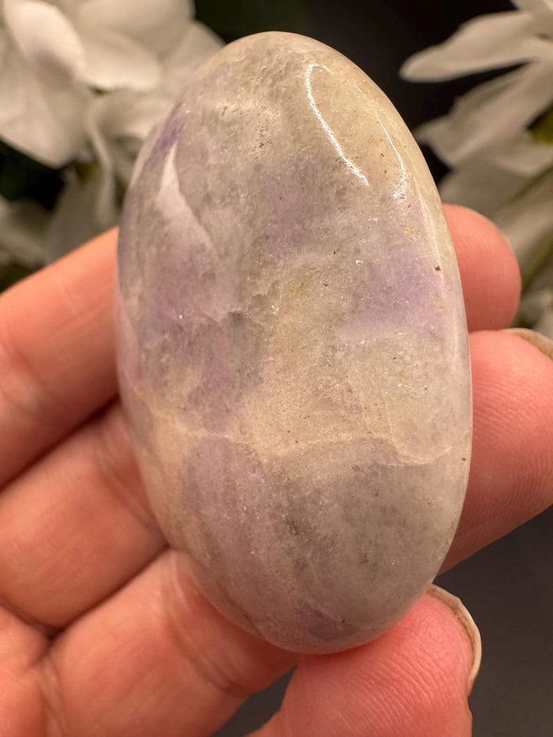 May include: Oval-shaped polished stone with a smooth, glossy surface. The stone features a blend of colors, including lavender, cream, and gray, with subtle variations in tone. The stone is held in a hand, suggesting its size and texture.