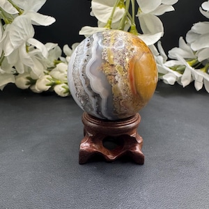 May include: A polished agate sphere with bands of gray, white, and brown hues, displayed on a carved wooden stand. The sphere's surface has a mix of textures and colors. White flowers are in the background.
