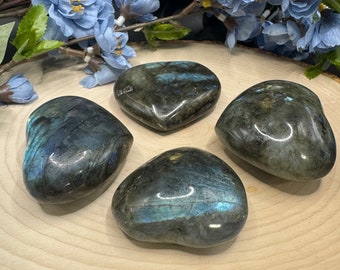 May include: Four heart-shaped labradorite crystal stones on a wooden surface. The stones are a dark gray color with iridescent blue and green flashes.