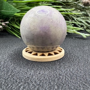 May include: A light purple sphere, possibly a decorative stone, sits on a small, round, light brown wooden stand. The sphere has a smooth surface with subtle color variations. The stand has a geometric design. The background includes greenery.