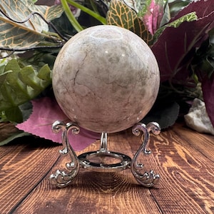 May include: A white and gray marble sphere on a silver stand with ornate scrollwork. The sphere is smooth and has a natural, polished finish.