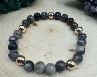 May include: A black and gray agate beaded bracelet with gold accents. The bracelet is made with round beads and is strung on elastic.
