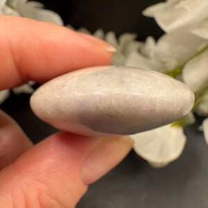 May include: A smooth, oval-shaped, light purple stone held between fingers. The stone has a polished surface with subtle color variations. The background features white flowers and a dark surface.