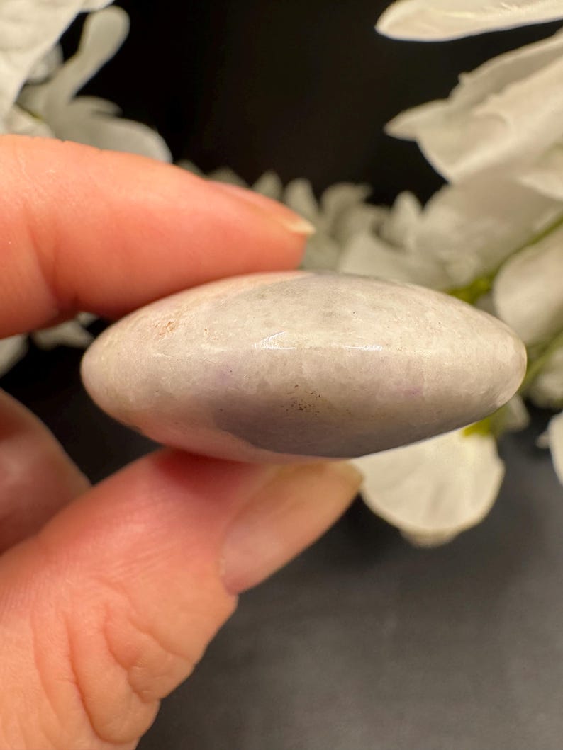 May include: A smooth, oval-shaped, light purple stone held between fingers. The stone has a polished surface with subtle color variations. The background features white flowers and a dark surface.