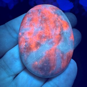 May include: An oval-shaped, polished stone with a vibrant, glowing orange and red pattern. The stone's surface has a marbled appearance with streaks of lighter gray and purple hues. The stone is held in a hand, with a dark blue background.