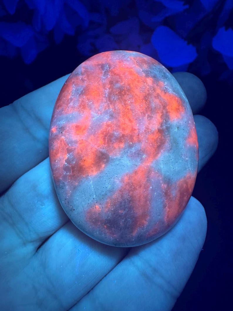 May include: An oval-shaped, polished stone with a vibrant, glowing orange and red pattern. The stone's surface has a marbled appearance with streaks of lighter gray and purple hues. The stone is held in a hand, with a dark blue background.