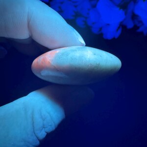 May include: A smooth, oval-shaped stone with a light gray base and orange and white accents. The stone is held between two fingers against a dark blue background, with blue floral shapes in the background.