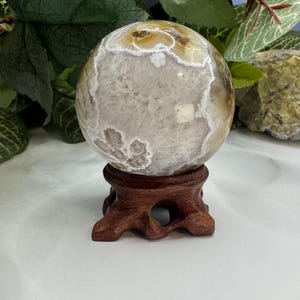 May include: A white agate sphere with brown and white banding, sitting on a wooden stand. The sphere is polished and has a smooth surface.