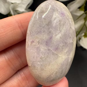 May include: A smooth, oval-shaped stone with a polished surface. The stone features a blend of pale lavender, cream, and gray hues, with subtle veining and variations in color. The stone is held in a hand, showcasing its natural beauty and texture.