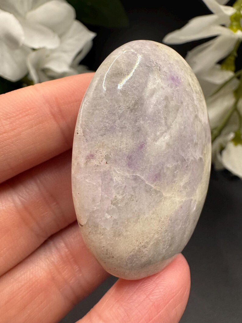 May include: A smooth, oval-shaped stone with a polished surface. The stone features a blend of pale lavender, cream, and gray hues, with subtle veining and variations in color. The stone is held in a hand, showcasing its natural beauty and texture.