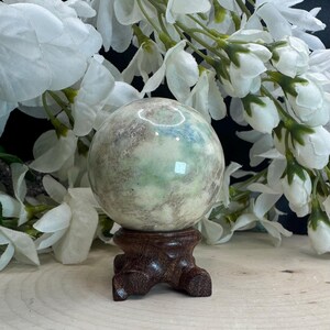 May include: A polished, spherical gemstone with a mottled appearance of green, gray, and cream colors. The sphere rests on a dark brown, carved wooden stand. White flowers with green stems are in the background.
