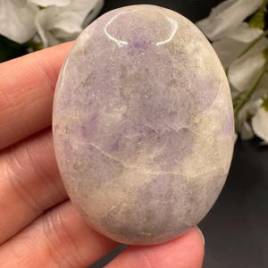 May include: An oval-shaped, polished stone with a blend of lavender, gray, and cream hues. The stone's surface has a smooth, glossy finish, and it is held in a hand. The background features white flowers and a dark surface.