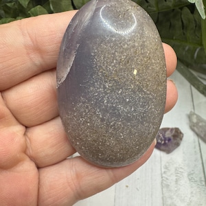 May include: A smooth, oval-shaped, light purple agate stone with a white circle pattern and brown flecks.