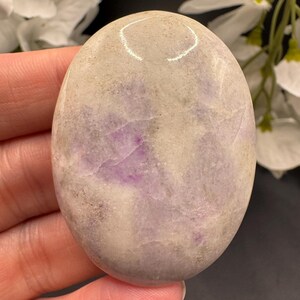 May include: Oval-shaped polished stone with a mix of lavender and cream colors. The stone has a smooth, glossy surface and a natural, organic appearance. The stone is held in a hand.