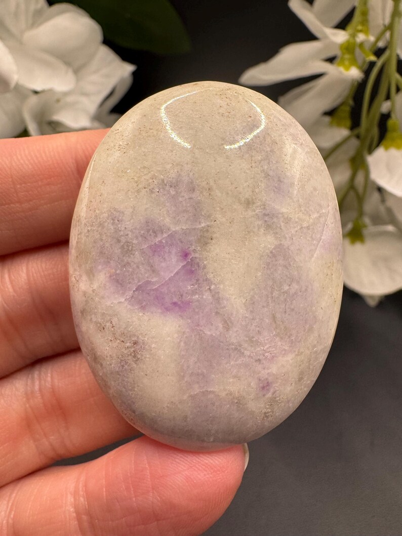 May include: Oval-shaped polished stone with a mix of lavender and cream colors. The stone has a smooth, glossy surface and a natural, organic appearance. The stone is held in a hand.