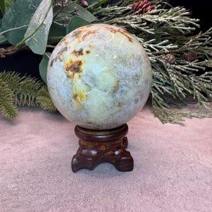 May include: A polished, spherical stone with a mottled surface featuring shades of pale green, cream, and brown mineral inclusions. The sphere rests securely on a dark, carved wooden tripod stand, showcasing a smooth, rounded texture throughout.