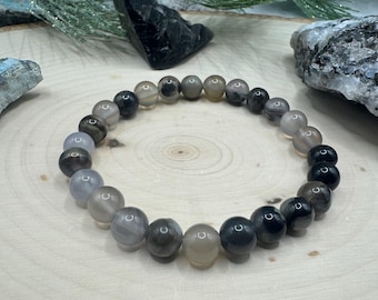May include: A gray and black beaded bracelet with round beads. The beads are graduated in size, with the largest beads at the center of the bracelet.