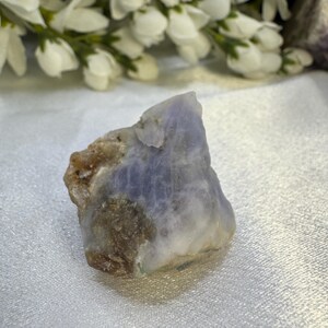 May include: A rough-cut, natural gemstone with a blend of blue, white, and brown hues. The stone has a textured surface and irregular shape. White flowers are in the background.