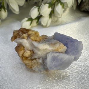 May include: A natural mineral specimen featuring a cluster of light blue, tan, and white crystals. The crystals have a rough, textured surface and are set against a soft, white background. The specimen is a unique piece of natural art.