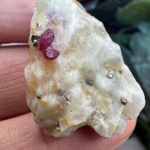 May include: A raw, irregular mineral specimen featuring a translucent white and pale yellow quartz matrix. Embedded within the stone are small, vibrant magenta-red crystal clusters and several tiny, metallic gold-colored pyrite flecks.