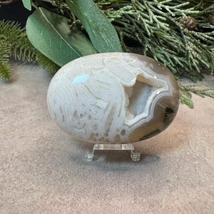 May include: An oval-shaped agate stone featuring intricate white and beige banding patterns with a central geode cavity filled with small, sparkling crystals. The stone is displayed on a small, clear acrylic stand for stability and viewing.