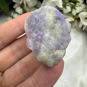 May include: A rough-cut, natural stone with a blend of lavender and pale gray hues. The stone has a textured surface and irregular shape, showcasing its raw, unpolished state. The background features white flowers.