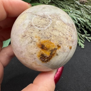 May include: A spherical, off-white stone sphere with brown and yellow markings. The surface has a cracked appearance. The sphere is held in a hand with red nail polish. Decorative greenery is in the background.