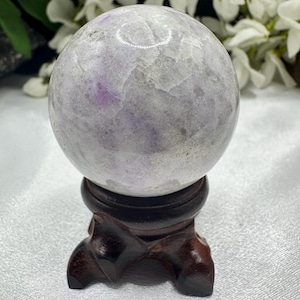 May include: A light gray crystal sphere with purple veining, displayed on a dark brown wooden stand. The sphere has a polished, smooth surface. The stand has a three-legged design. The background features white flowers.