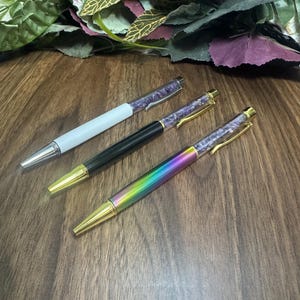 May include: Three ballpoint pens with gold accents. One pen is white, one is black, and the third has a rainbow finish. All three pens have a purple, crystal-like design on the top portion.