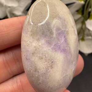 May include: Oval-shaped polished stone with a blend of colors. The stone features a base of light beige and gray hues, with streaks and patches of lavender and purple. The smooth surface reflects light, highlighting the natural patterns.