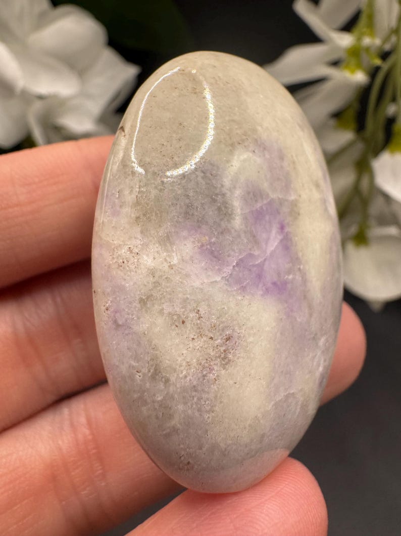 May include: Oval-shaped polished stone with a blend of colors. The stone features a base of light beige and gray hues, with streaks and patches of lavender and purple. The smooth surface reflects light, highlighting the natural patterns.