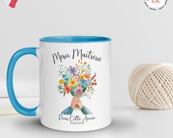 May include: White ceramic mug with a blue rim and handle. The mug has a floral design with the text "Merci Maitresse Pour Cette Annee 2023/2024".