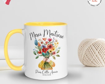 May include: White ceramic mug with a yellow rim and handle. The mug has a floral design with a bouquet of colorful flowers held by a hand wearing a yellow sweater. The text "Merci Maitresse Pour Cette Annee 2023/2024" is printed on the mug.