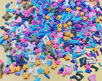 May include: A colorful assortment of sprinkles in various shapes, including stars, hearts, musical notes, and airplanes. The sprinkles are in shades of pink, blue, purple, white, and gold. These decorative sprinkles are perfect for decorating baked goods.