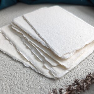 May include: A stack of white handmade paper sheets with deckled edges. The paper has a textured surface and is perfect for writing, drawing, or crafting.
