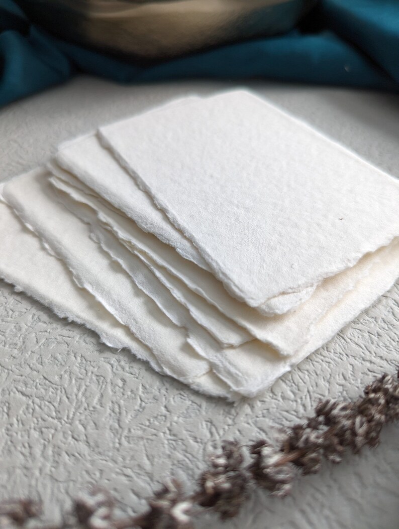 May include: A stack of white handmade paper sheets with deckled edges. The paper has a textured surface and is perfect for writing, drawing, or crafting.