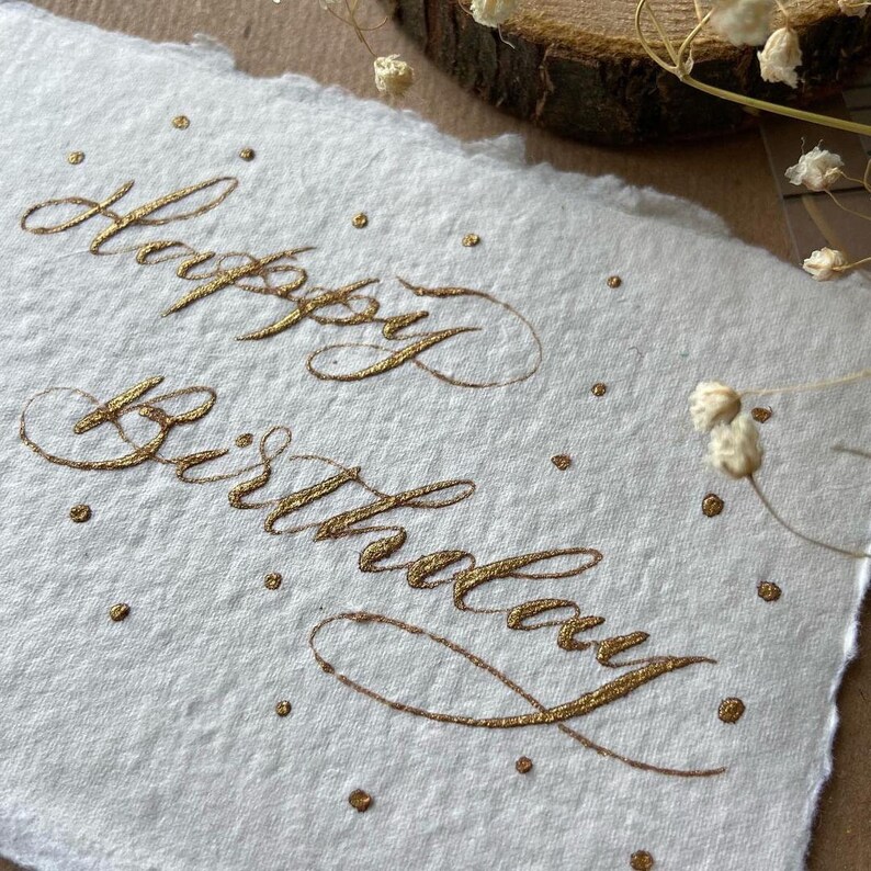 May include: A white card with the words "Happy Birthday" written in gold calligraphy. The card is decorated with small gold dots.
