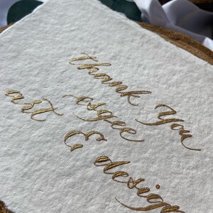 May include: A white card with the words "Thank you" and "for the design" written in gold calligraphy.