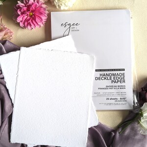 May include: A pack of 25 sheets of handmade, 100% cotton rag, acid-free, deckle edge paper. The paper is white and has a textured surface. The pack is labeled "Handmade Deckle Edge Paper" and includes the text "Papier au bords franges fait a la main".