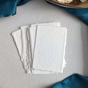 May include: A stack of five white, textured, handmade paper cards with deckled edges. The cards are on a light gray textured surface with a teal blue fabric in the background.